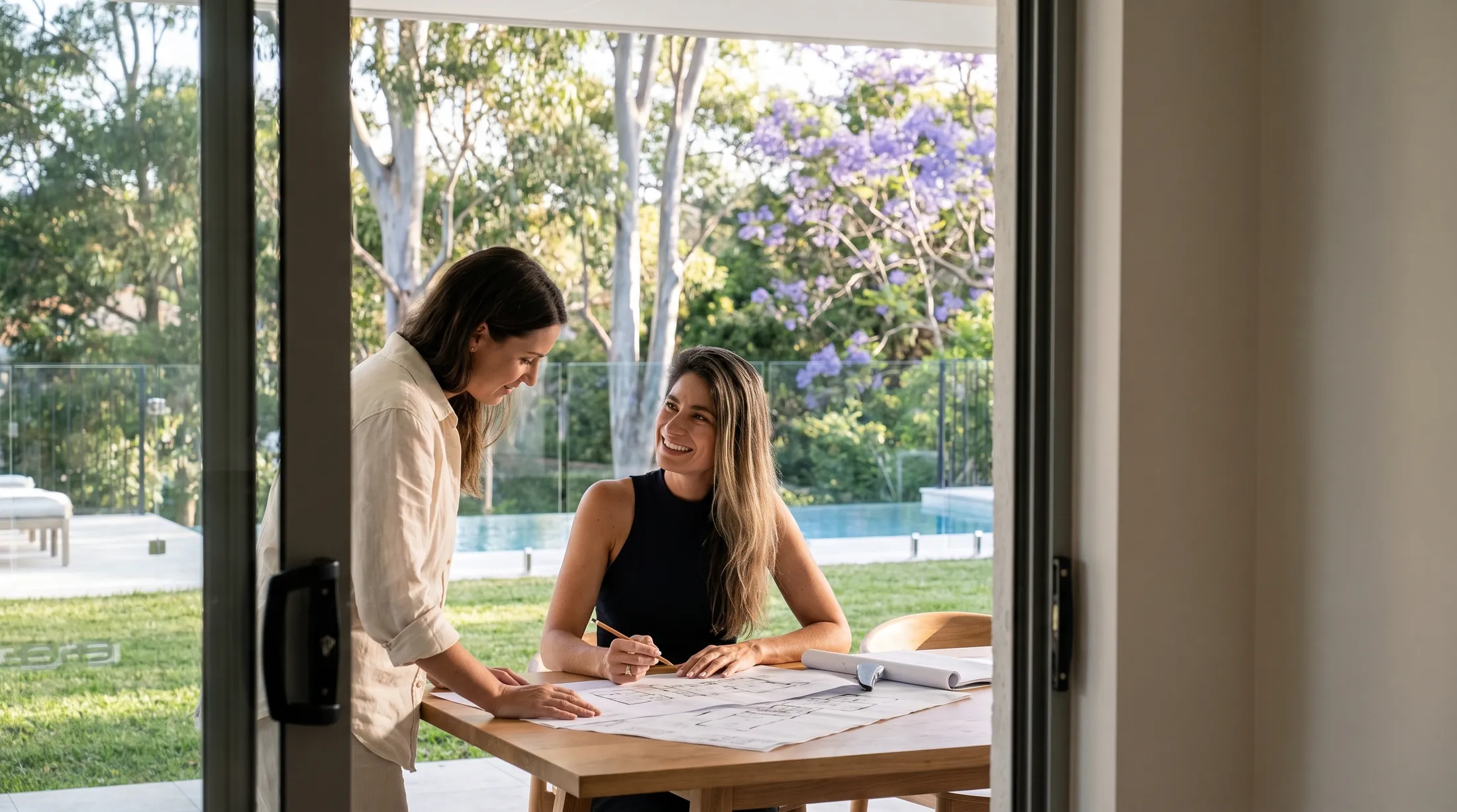 Monica Vogel seated at an outdoor table with a client, going through a set of architectural drawings, a pool and garden behind them.